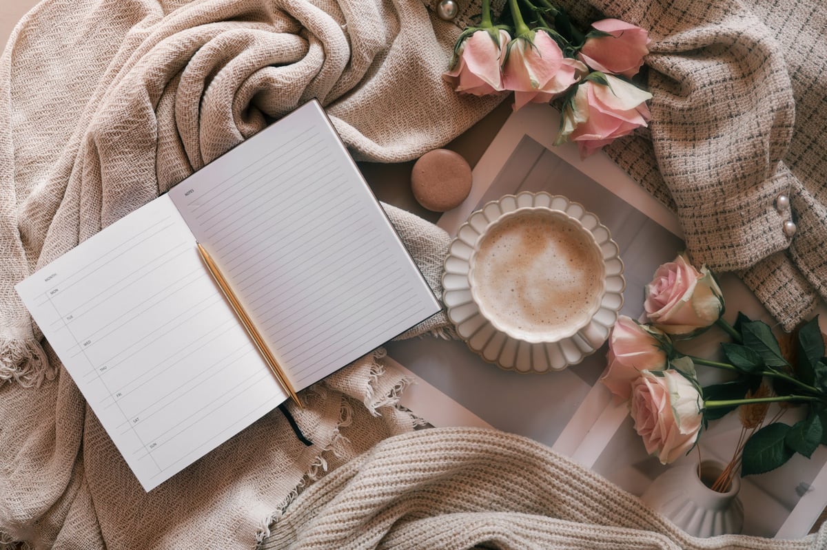 Flat lay of an open weekly planner with a pen, coffee cup, pink roses, and cozy neutral fabrics.
